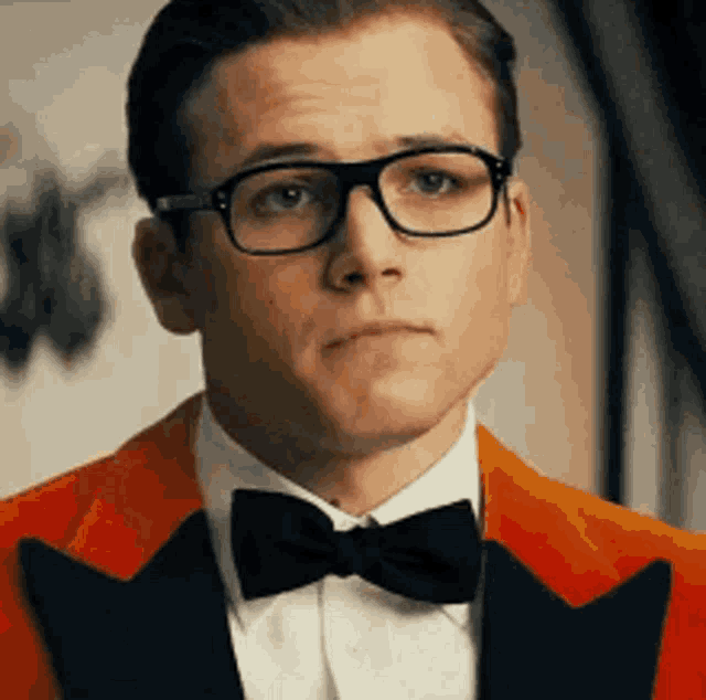 a man wearing glasses , a red suit and a black bow tie is making a funny face .