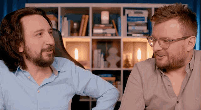 two men are talking in front of a bookshelf and one has glasses on