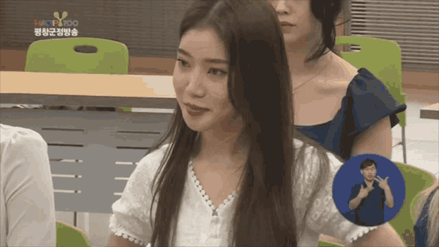 a woman in a white shirt sits in a classroom with a sign language sign behind her