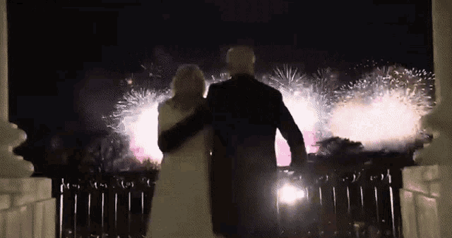 a man and a woman are standing on a balcony looking at fireworks