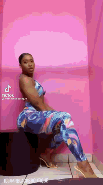 a woman is sitting on a stool in front of a pink wall wearing a blue and pink outfit .
