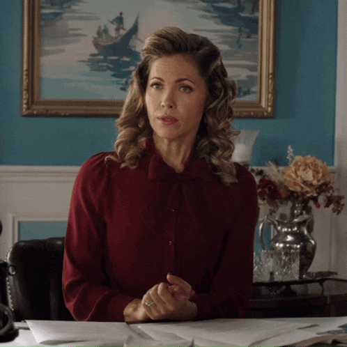 a woman in a red shirt sits at a desk in front of a painting
