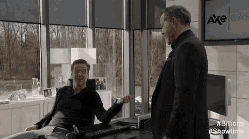 a man sitting at a desk talking to another man standing in front of a screen that says axe