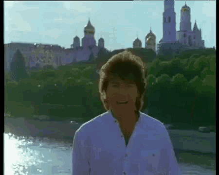 a man in a white shirt is standing in front of a body of water with a castle in the background .