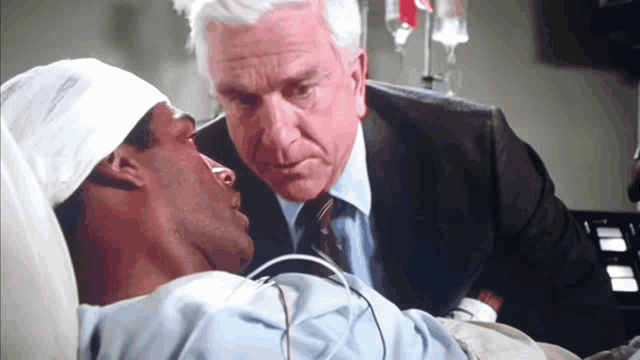 a man in a suit stands next to a man in a hospital bed with a bandaged head