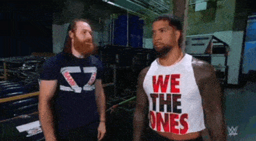two men are standing next to each other in a room and one of them is wearing a we the ones tank top .