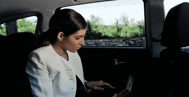 a woman in a white jacket sits in the back seat of a car using a tablet