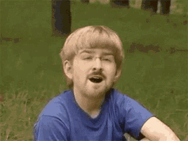 a young man with glasses and a mullet is sitting in the grass making a funny face .