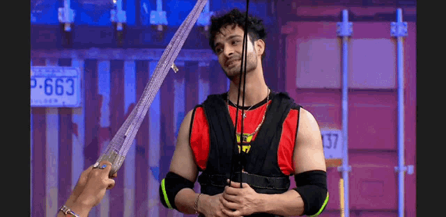 a man in a red shirt and black vest is holding a sword .