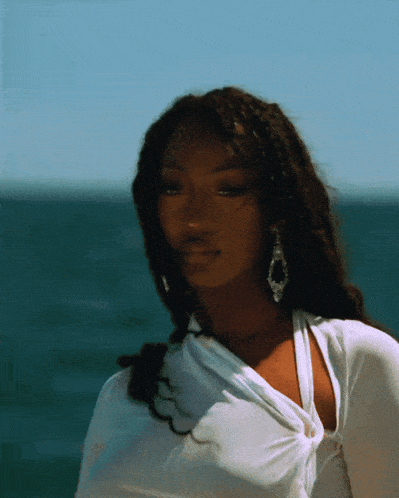 a woman wearing earrings and a white shirt stands in front of the ocean