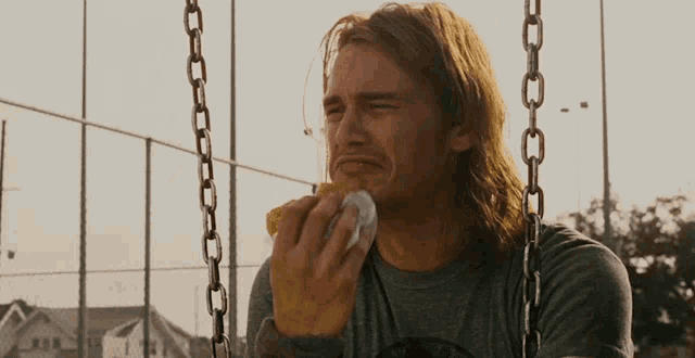 a man with long hair is sitting on a swing eating a piece of paper