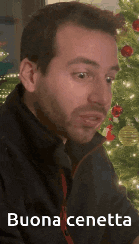 a man sitting in front of a christmas tree with the words buona cenetta on the bottom