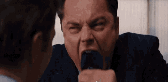 a man in a suit is screaming into a microphone while another man watches .