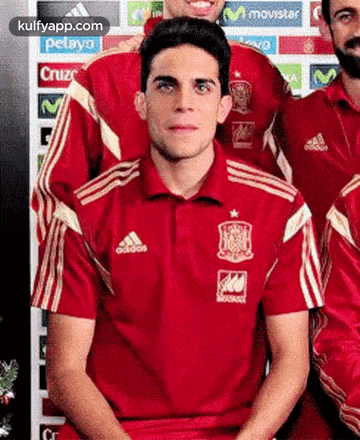 a man in a red adidas shirt is sitting in front of a group of other men .