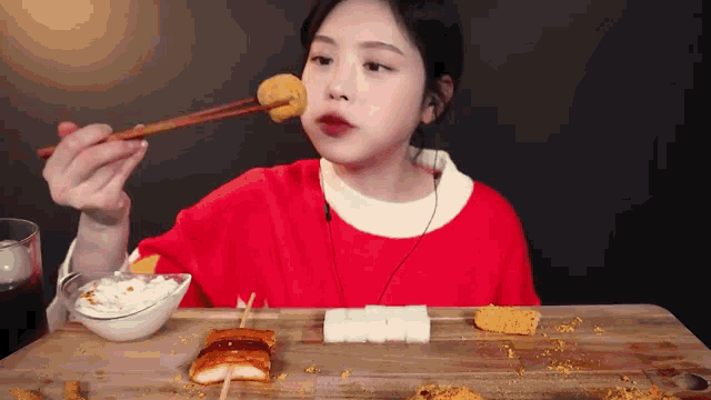 a woman in a red shirt is eating a skewer of food with chopsticks .