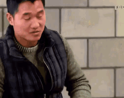 a man wearing a vest and sweater is standing in front of a brick wall with his eyes closed