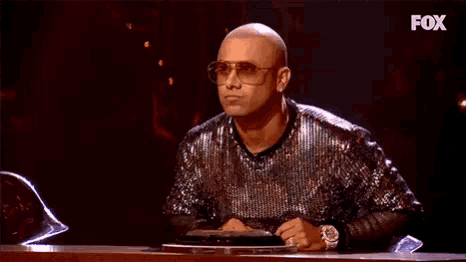 a bald man wearing sunglasses and a sequined shirt is sitting at a table with a fox logo behind him .