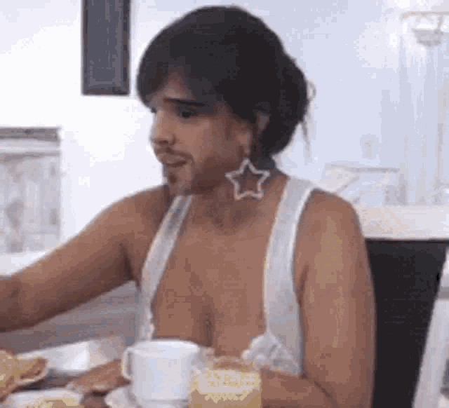 a man without a shirt is sitting at a table with a cup of coffee and a plate of food .