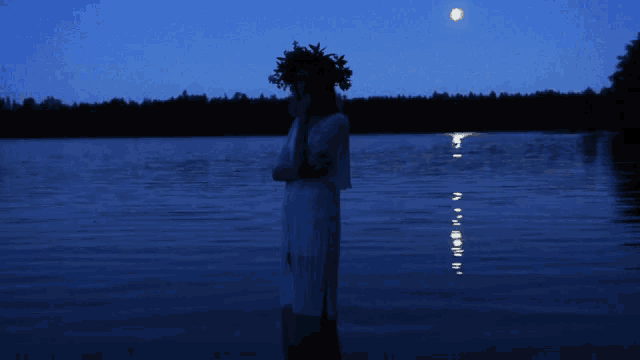 a woman with a wreath on her head stands in front of a lake at night