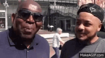 two men wearing sunglasses and a hat are standing next to each other on a street .