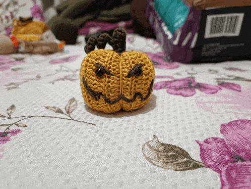 a crocheted pumpkin sits on a bed with purple and white flowers