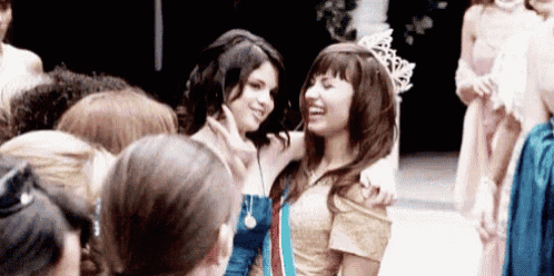 two women are standing next to each other in a crowd of people . one of the women is wearing a crown .