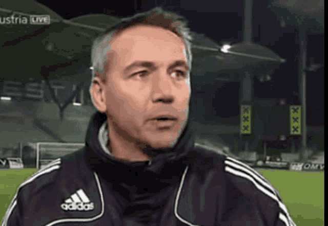 a man wearing an adidas jacket stands in front of a soccer field
