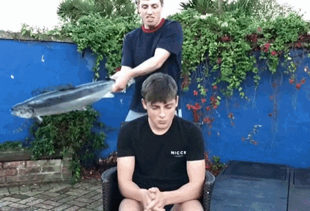 a man is holding a fish over another man 's head who is wearing a nicc t-shirt