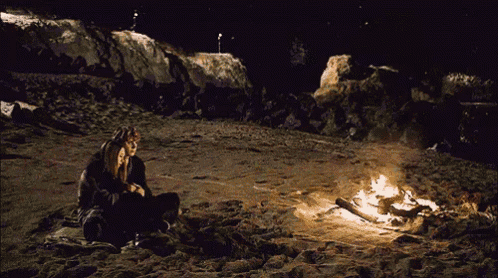 a man and a woman sit by a fire on a beach at night