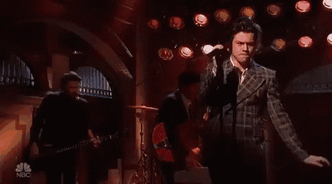 a man in a plaid suit is singing into a microphone on a stage while a band plays behind him .