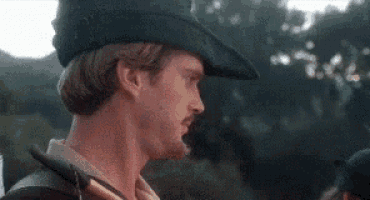 a man with a beard wearing a hat and holding a sword is standing in the woods .