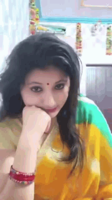 a woman in a yellow and green saree looks at the camera with her hand on her chin