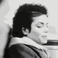 a black and white photo of a man with curly hair sitting next to a wall .