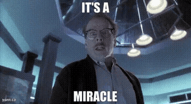 a man with glasses is standing in a dark room with the words `` it 's a miracle '' written on the screen .