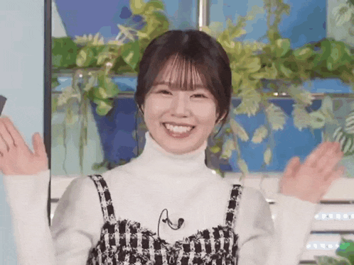 a woman in a white turtleneck sweater is smiling and waving her hands in front of a window .