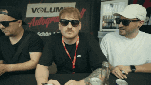 three men are sitting at a table in front of a sign that says volum autograph