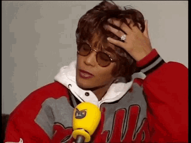 a woman wearing sunglasses and a red jacket is holding her head while talking into a yellow microphone .