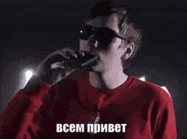 a man wearing sunglasses and a red shirt is singing into a microphone in russian .