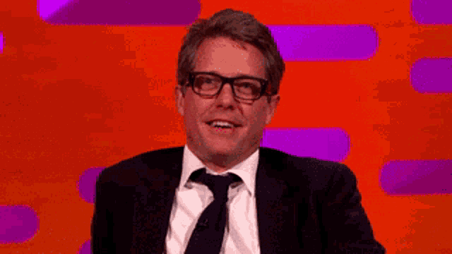 a man wearing glasses and a suit is sitting in front of an orange background