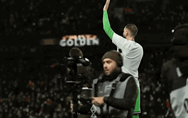 a man in a white shirt and green shorts stands in front of a camera and a scoreboard that says outro