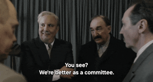 a group of men are sitting in a room and one of them is asking if they are better as a committee