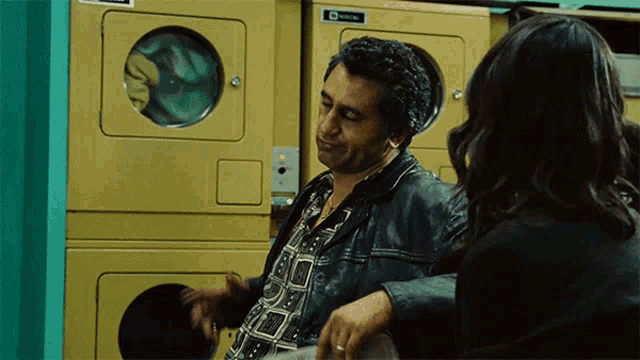 a man in a leather jacket talks to a woman in front of a stack of washing machines