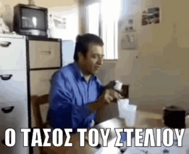 a man in a blue shirt is sitting at a table in front of a television and a fridge .