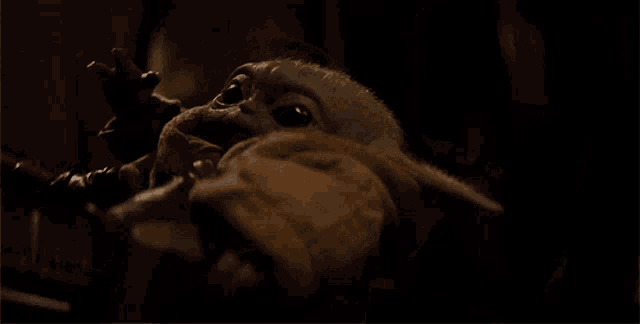 a baby yoda from star wars is sitting in a dark room and waving at the camera .