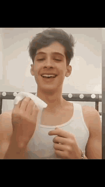 a young man in a white tank top is smiling and holding a piece of paper in his hand .