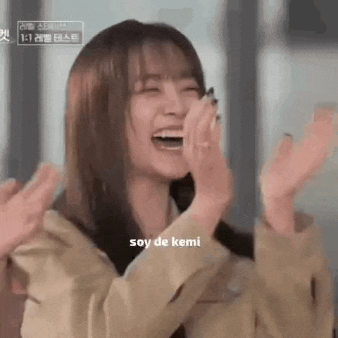 a woman is laughing and clapping her hands in the air while wearing a tan jacket .