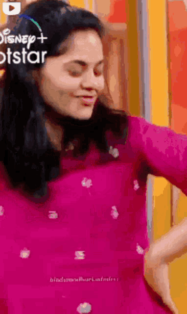 a woman in a pink shirt is dancing in front of a sign that says disney plus hotstar
