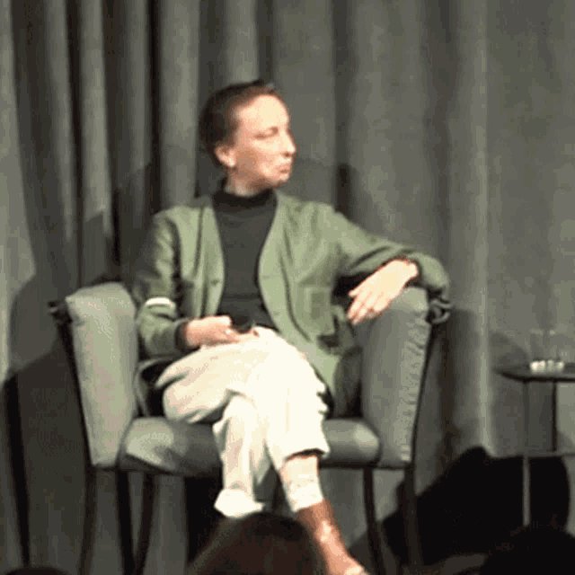 a woman in a green jacket is sitting in a chair with her legs crossed
