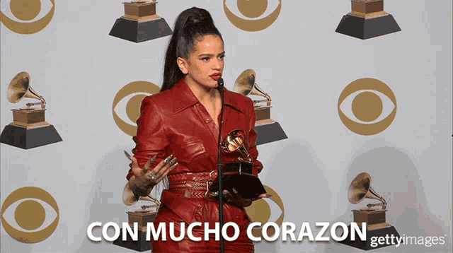 a woman in a red leather dress is holding a grammy in front of a wall that says con mucho corazon on it