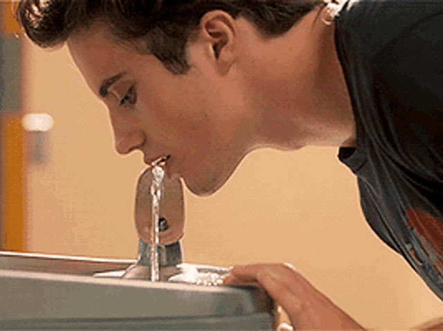 a man drinking water from a fountain in a bathroom